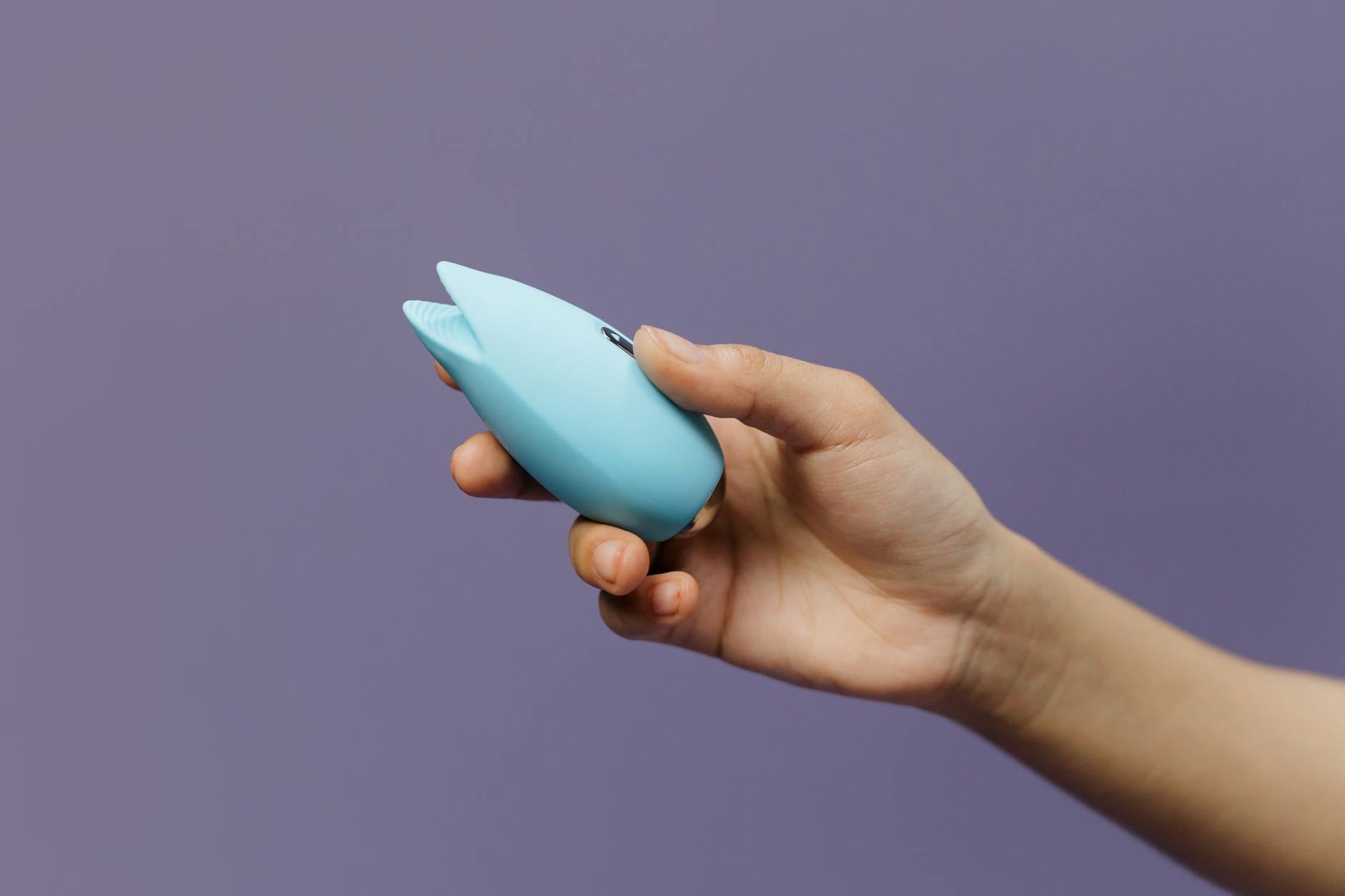Blue silicone clitoral vibrator held in open palm against solid purple background